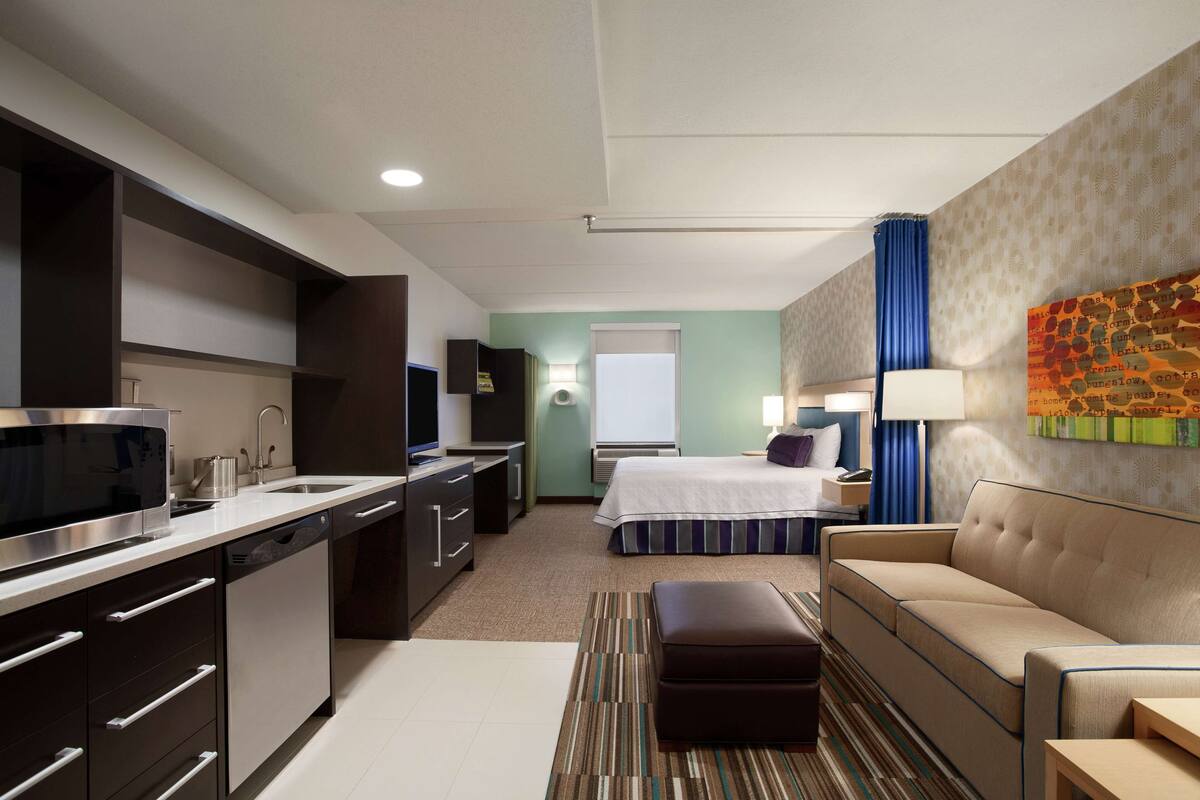 A compact hotel room with a kitchenette, a bed with a striped bedspread, a sofa, a small table, and a wall-mounted artwork.