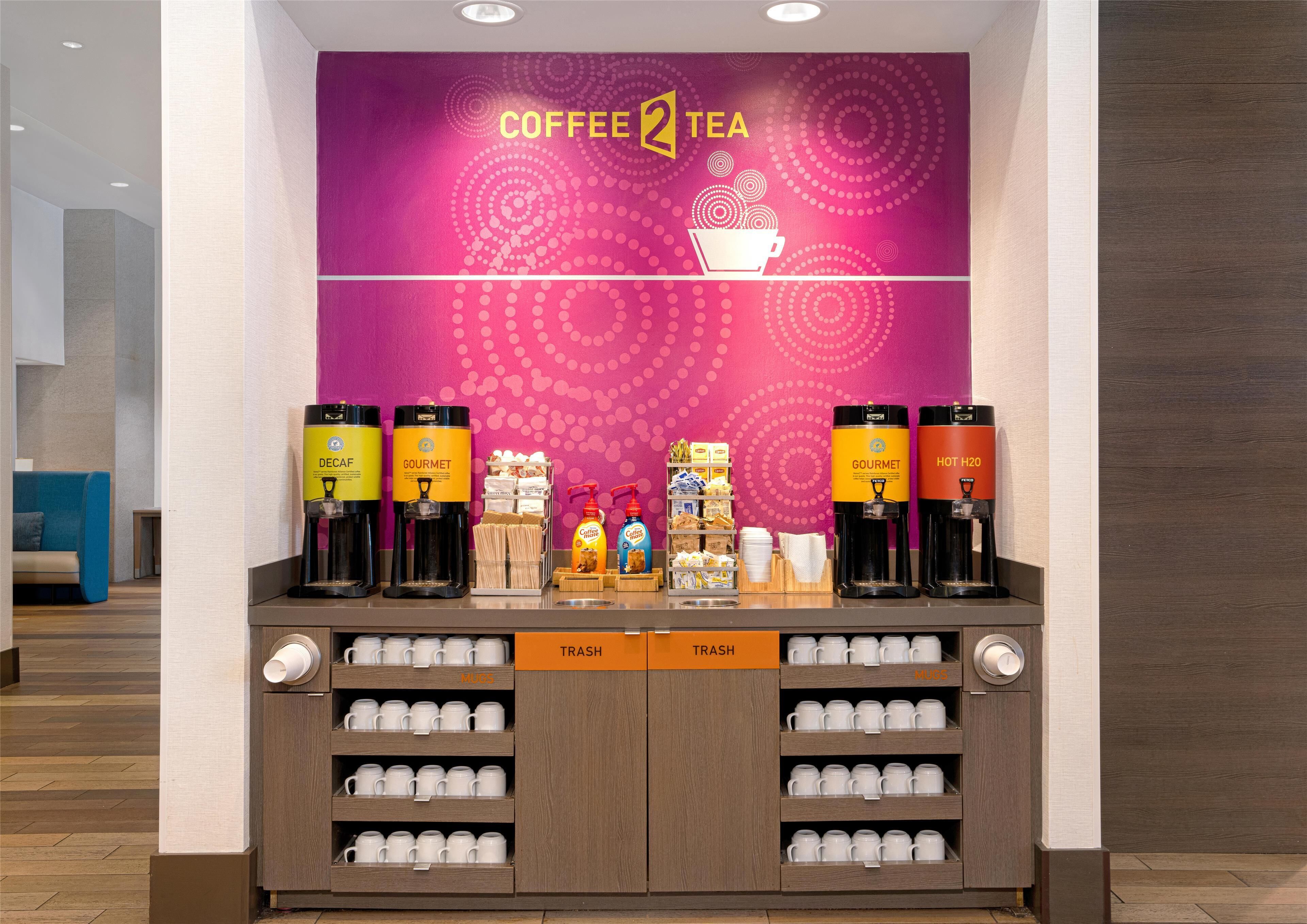 Cantinho com café e chá, máquinas de distribuição com a marca, alguns petiscos e uma lixeira.
