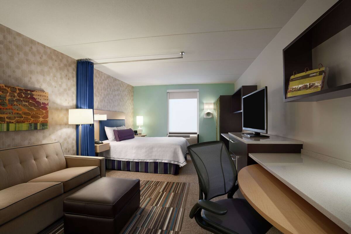 A hotel room with a bed, a sofa, a desk, a TV, and a window.