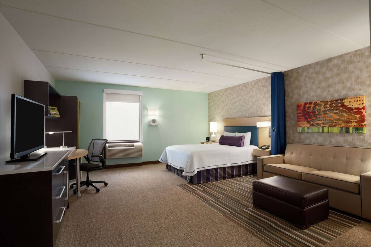 A hotel room with a bed, a desk with a computer, a TV, a sofa, and a decorative wall piece.