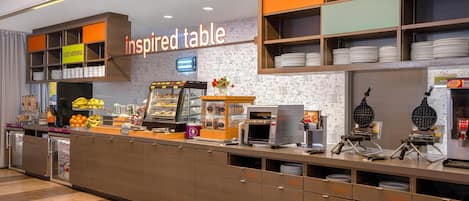 A breakfast bar with a waffle maker, a coffee machine, and a fruit display.