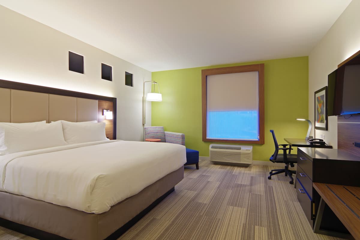A hotel room with a large bed, a desk, a chair, a lamp, and a large window.