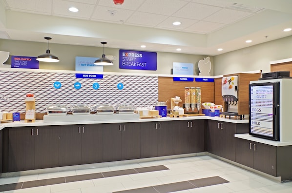 A modern breakfast area with a counter displaying hot food options, a digital menu board, and a fruit display.