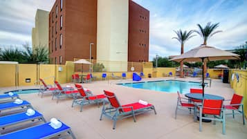 Área da piscina com espreguiçadeiras, piscina e um edifício ao fundo.