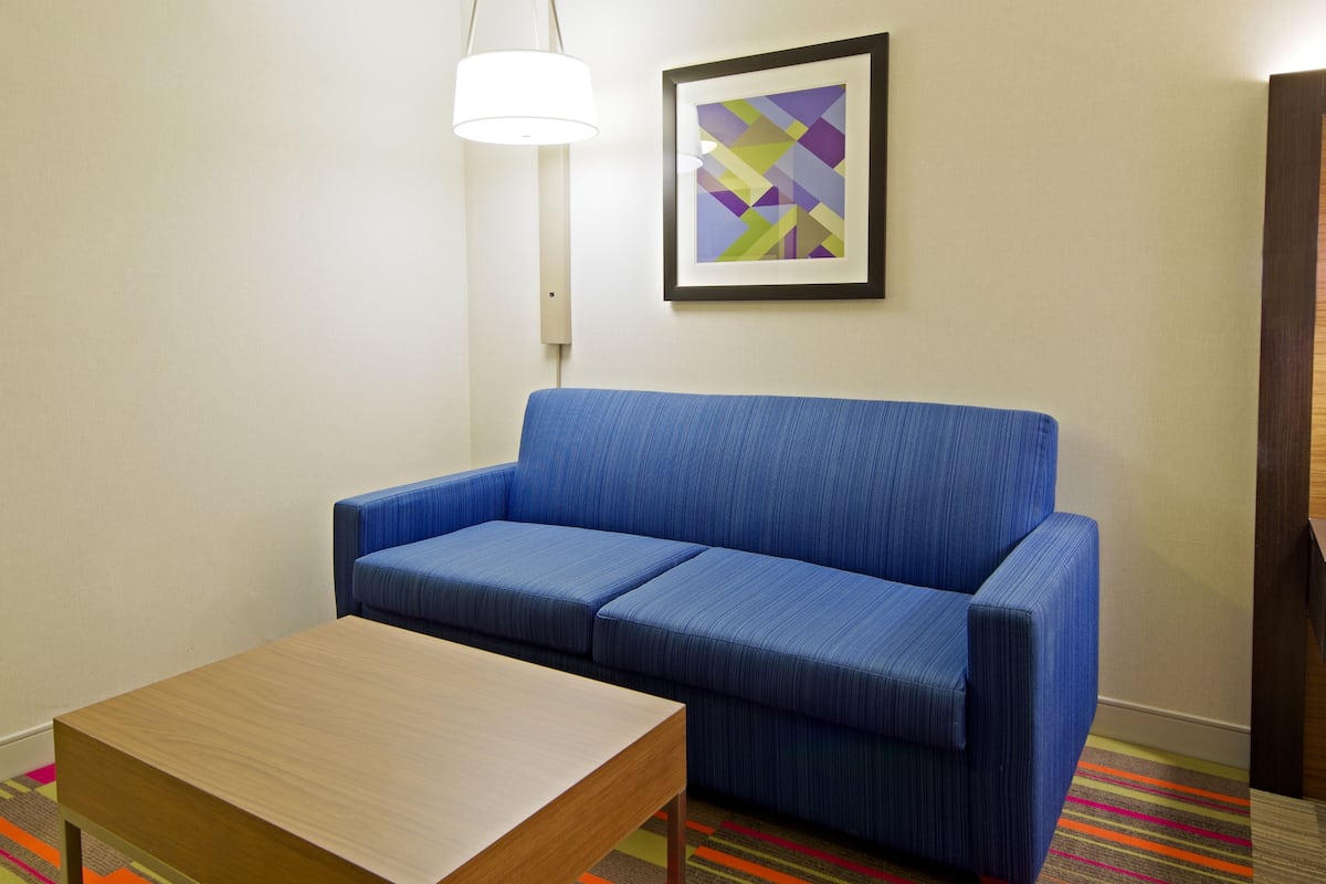 A room with a blue sofa, a wooden table, a colorful striped carpet, a white wall, and a framed abstract painting.