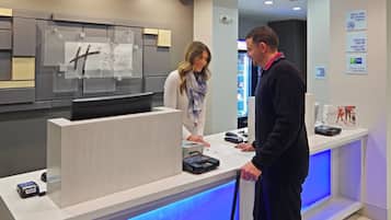 A hotel reception desk with a digital check-in kiosk, a security camera, and a sign indicating a 100% non-smoking property.