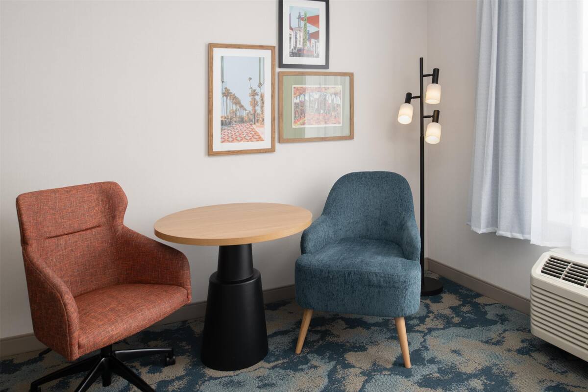 A room with a round table, two chairs, framed pictures on the wall, a floor lamp, and a window with curtains.
