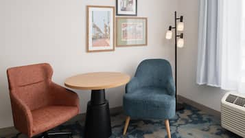 Una habitación con una mesa redonda, dos sillas, cuadros en la pared, una lámpara de pie y un ventanal con cortinas.