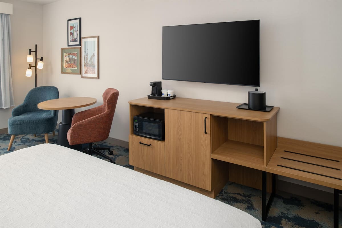 A hotel room with a bed, a desk with a microwave, a coffee maker, a TV, and two chairs.