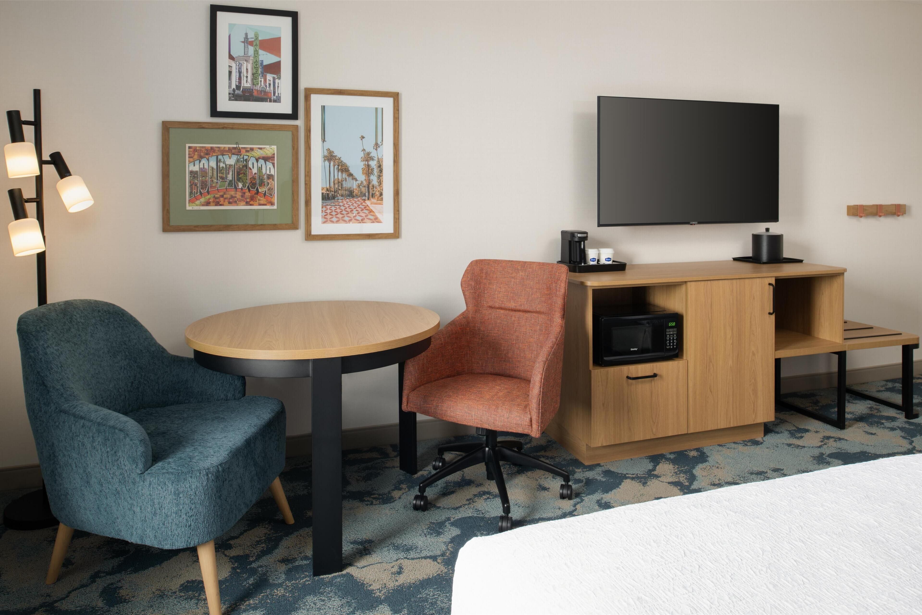 Habitación de hotel con una cama, un escritorio con televisor de pantalla plana, microondas, cafetera y dos sillas. En la pared, hay cuadros enmarcados.
