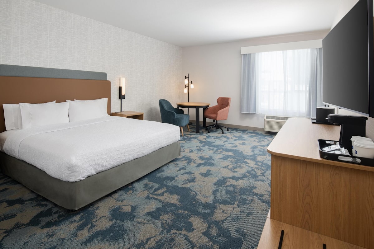 A hotel room with a large bed, a desk with a computer, two chairs, a small table, and a window with curtains.