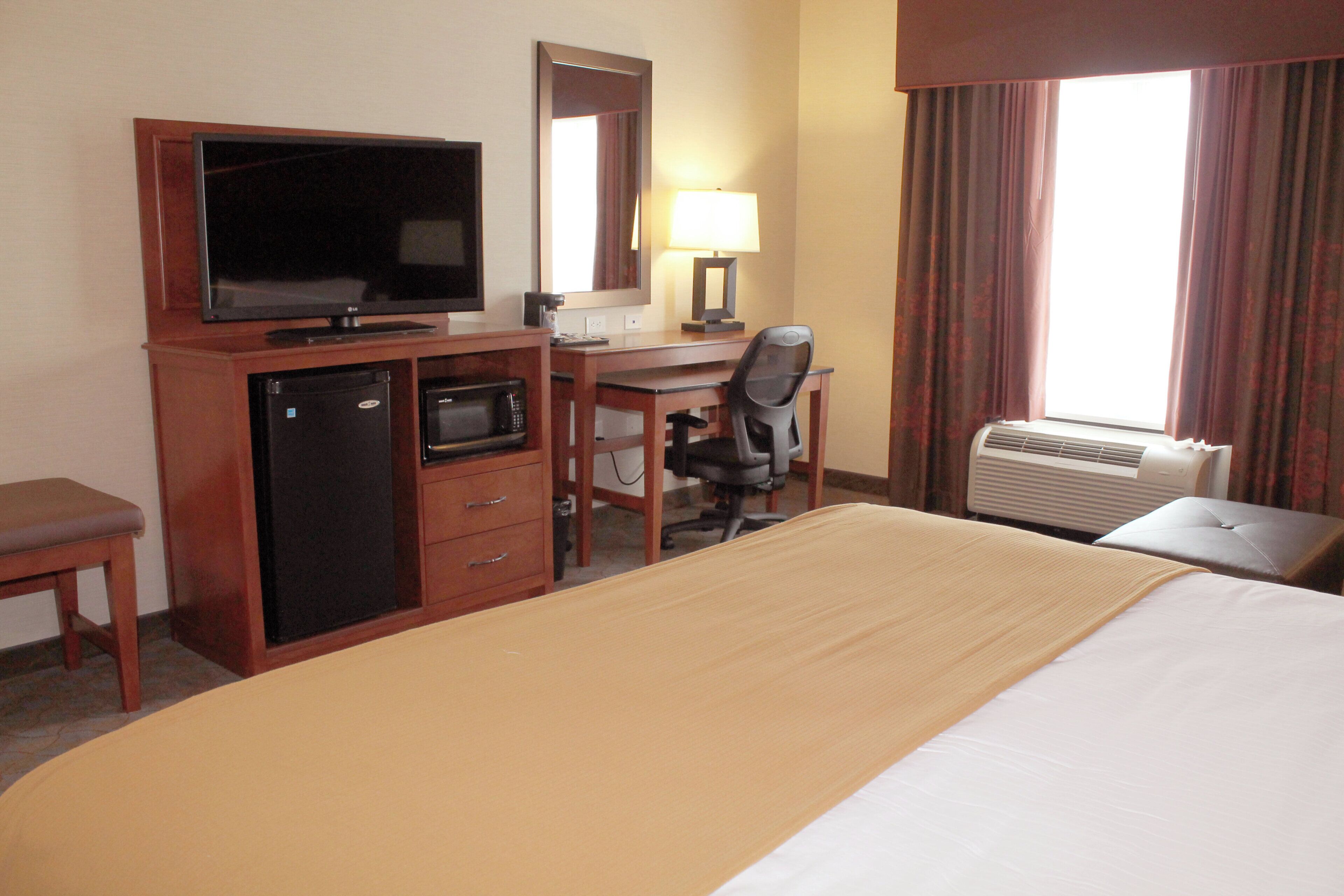 A hotel room with a bed, a desk, a television, a mini-fridge, a microwave, a chair, a lamp, and an air conditioning unit.