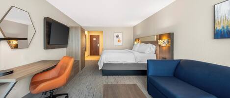 A hotel room with a bed, a desk, an orange office chair, a blue sofa, a TV mounted on the wall, and a painting.