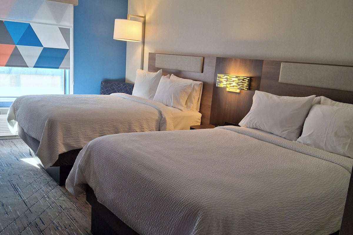 A hotel room with two beds, a large window with a geometric view, and a wall-mounted lamp.