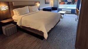 Una habitación de hotel moderna con una cama grande, mesitas de noche, un pequeño rincón de descanso y vistas a la ciudad a través del ventanal.