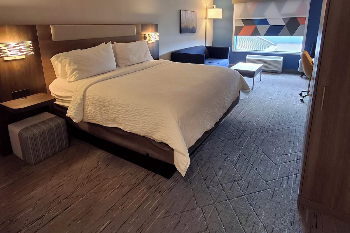 A modern hotel room with a large bed, bedside tables, a small seating area, and a view of the city through the window.