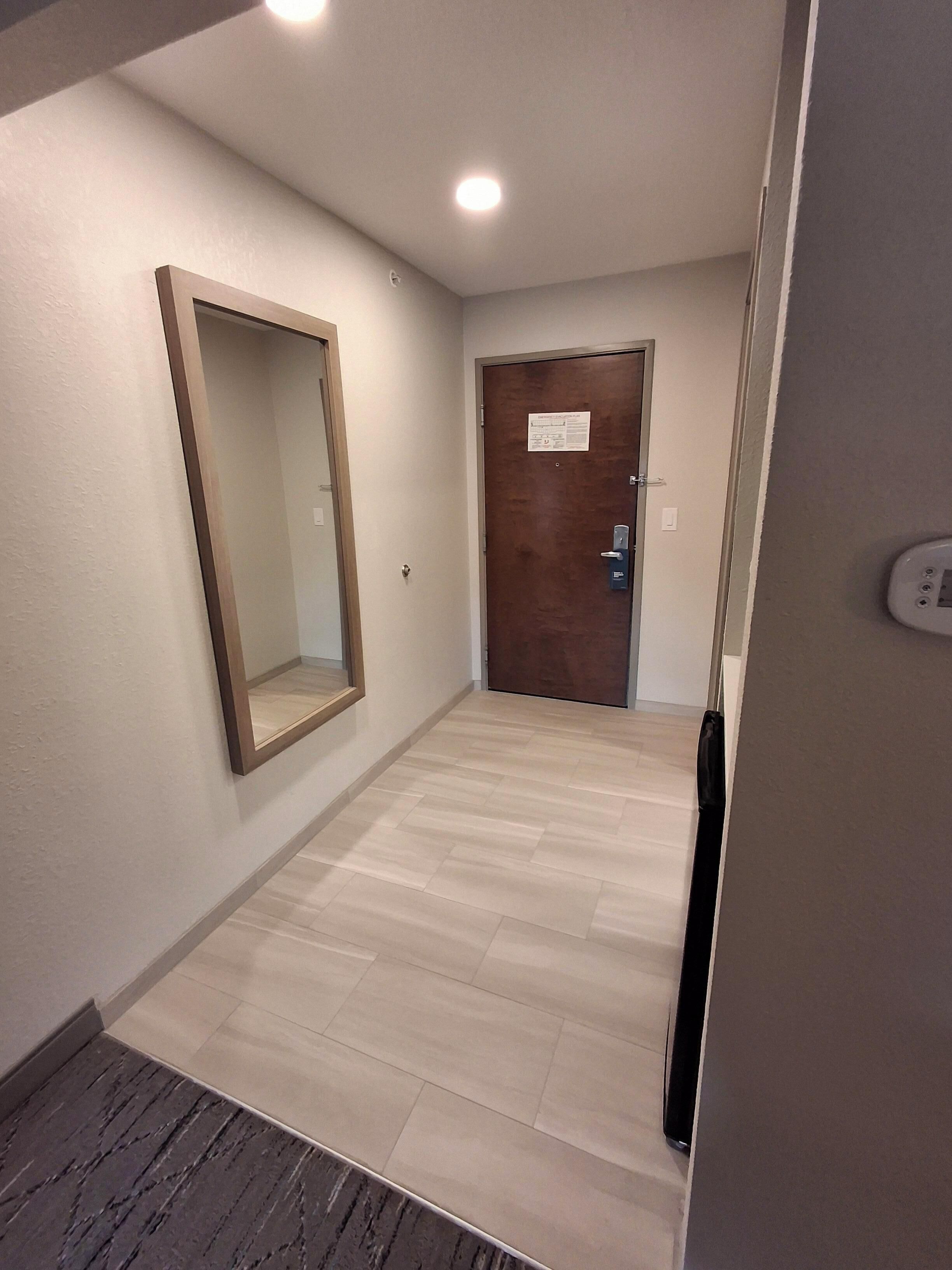 Un pasillo con una puerta de madera, un espejo y un interruptor de luz en la pared.