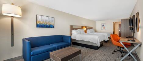 Habitación de hotel con sofá azul, mesa de centro de madera, cama con ropa blanca, sillón naranja y escritorio con lámpara.