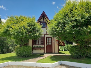 A quaint, single-story house with a steeply pitched roof, surrounded by lush greenery and a well-maintained lawn.