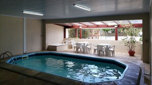 Una piscina interior con solárium azulejado, un porche cubierto con sillas y mesas de plástico blanco, y vistas al exterior a través de amplias ventanas.