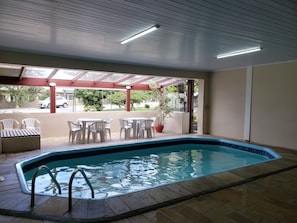 An indoor swimming pool with a tiled floor, surrounded by white plastic chairs and tables, under a glass roof with a view of the street outside.