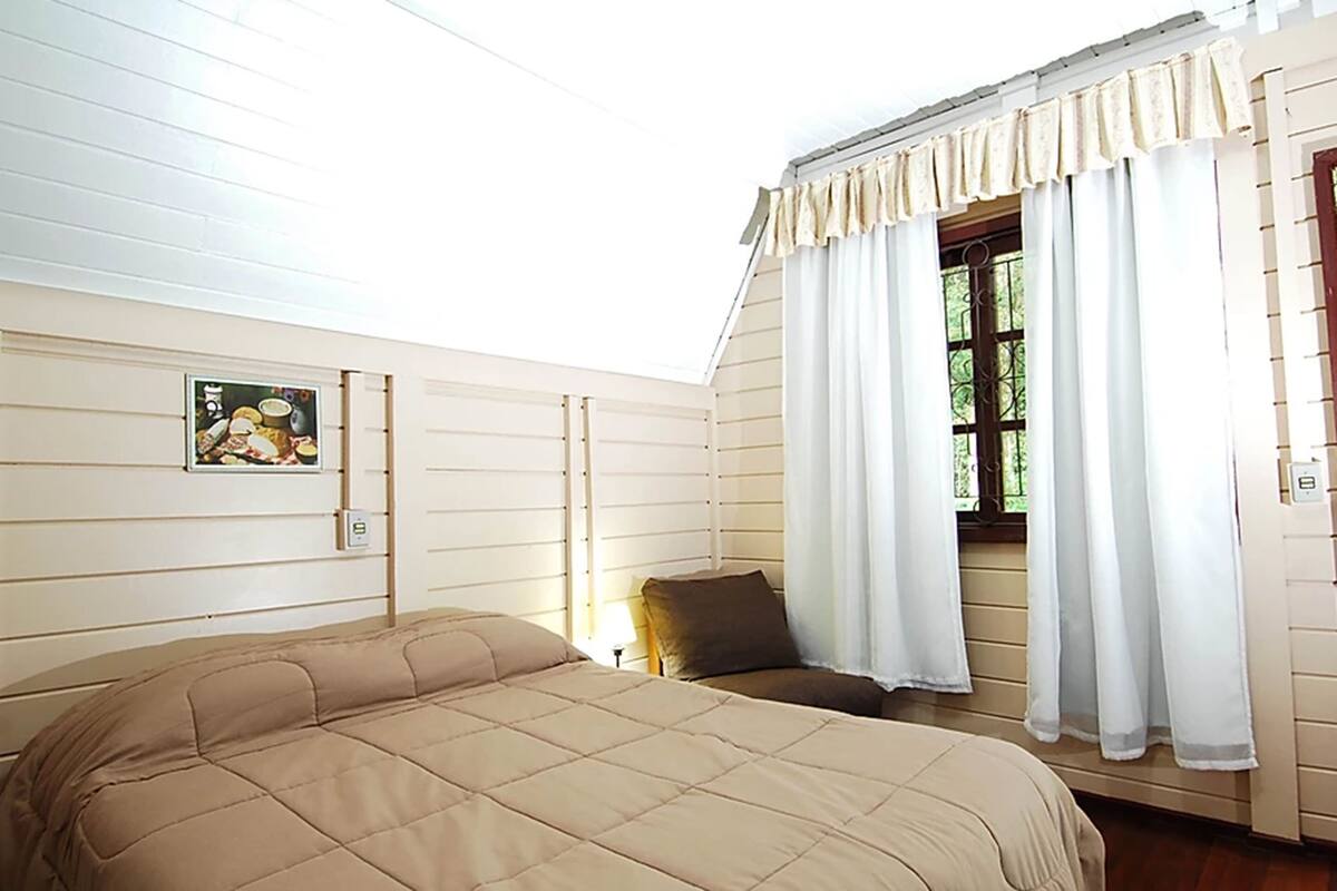 Un dormitorio con una cama grande, una ventana con cortinas, un cuadro colgado en la pared y una mesita de noche con lámpara.