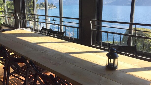 Een lange houten tafel met stoelen, een lantaarn en uitzicht op een meer met bergen op de achtergrond.
