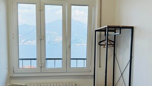 Een slaapkamer met een bed, een raam met uitzicht op zee en bergen, een radiator, een zwarte tafel en een metalen plank met een deken.