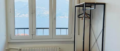 A bedroom with a bed, a window with a view of the sea and mountains, a radiator, a black table, and a metal shelf with a blanket.