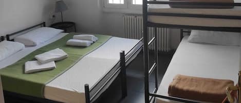 A dormitory room with bunk beds, a desk, a chair, a radiator, and a window with blinds.