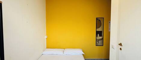 Una cama individual en una habitación con paredes amarillas, una puerta blanca y un espejo pequeño en la pared.