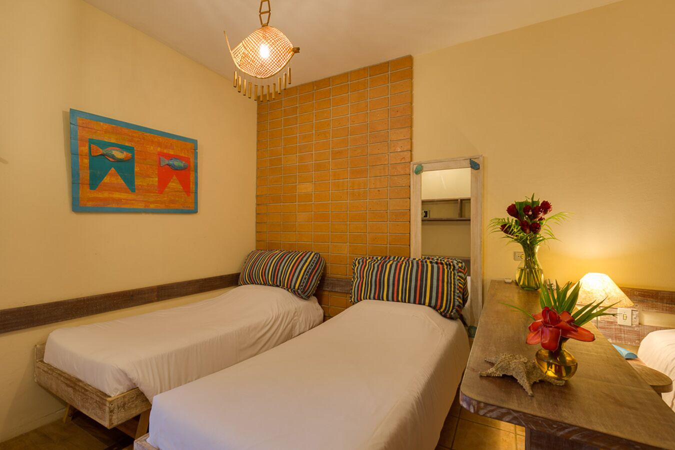 Habitación de hotel con dos camas individuales, pared de azulejos, un cuadro, un espejo y una mesita con un jarrón de flores.