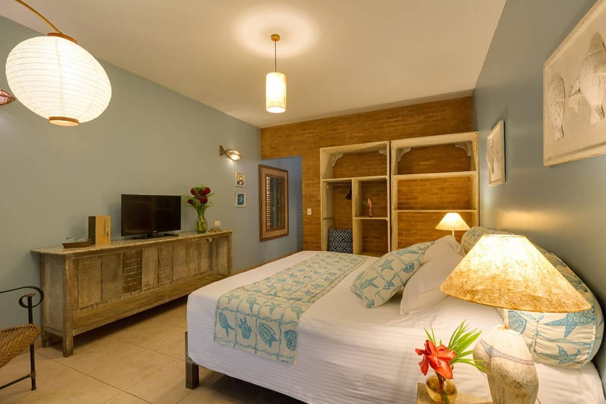 A bedroom with a bed, bedside lamp, TV, wooden dresser, and a wall-mounted shelf.