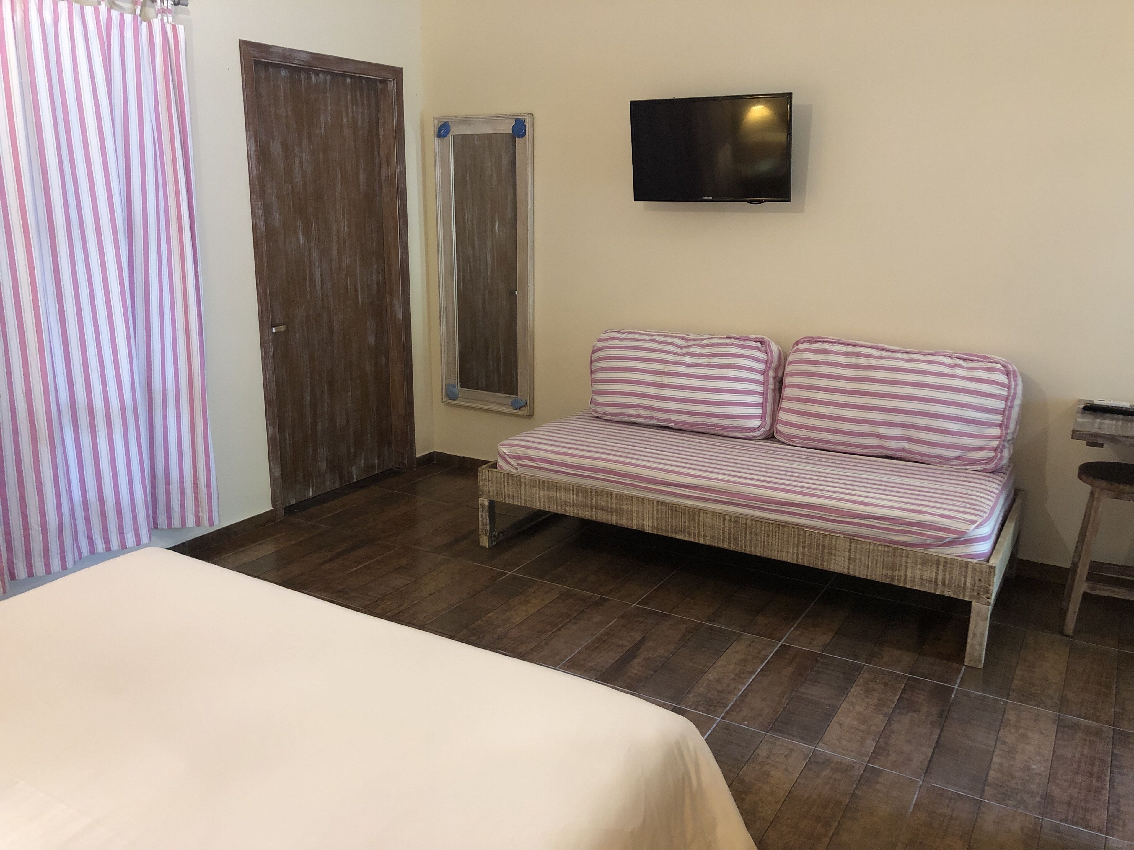 Una habitación con una cama, un sofá con cojines a rayas, un televisor montado en la pared y una puerta de madera.
