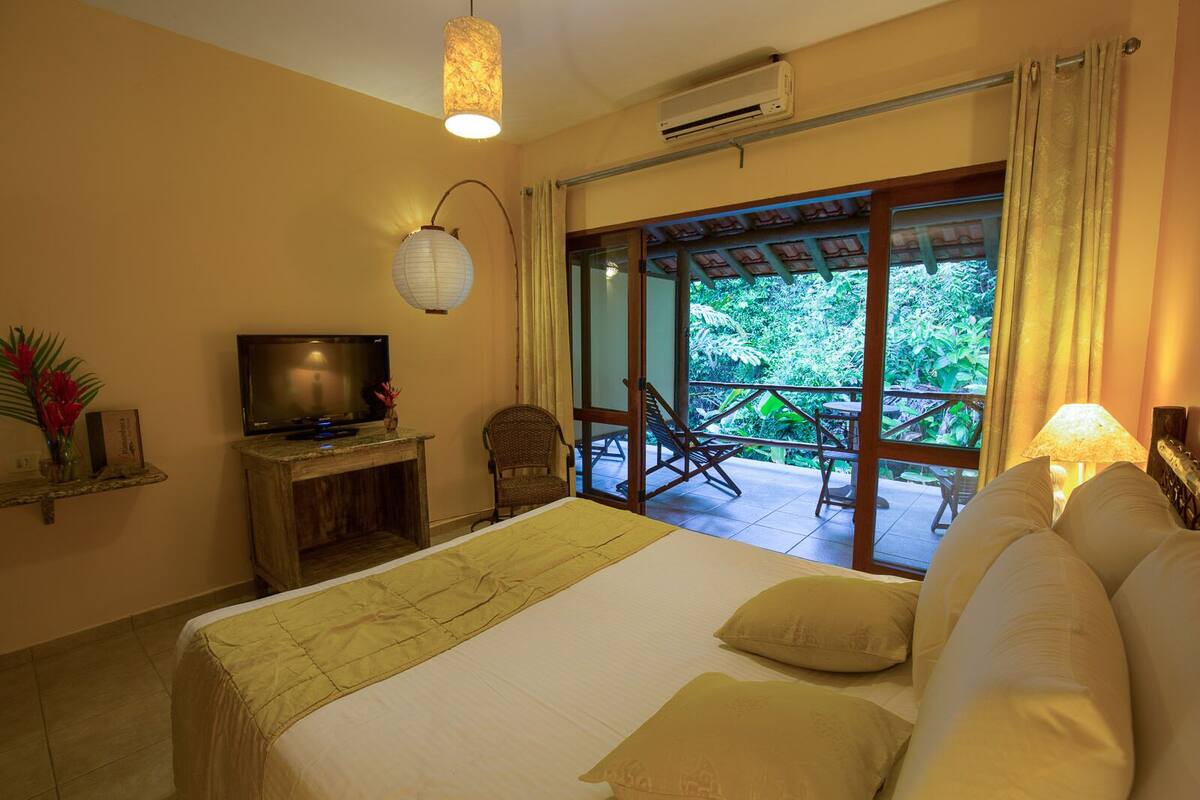 A hotel room with a bed, a television, a desk, a chair, and a balcony with a view of greenery.