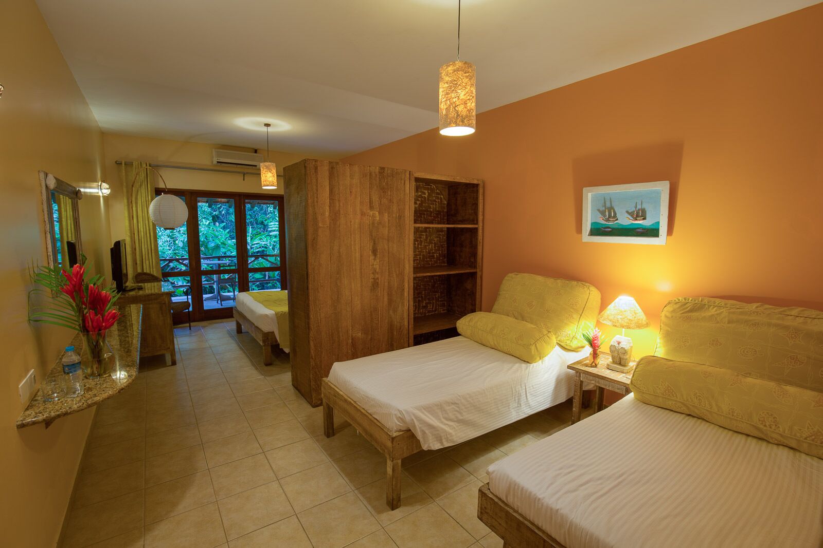 A hotel room with two beds, a wooden bookshelf, a dining area with a table and chairs, and a large window with a view of greenery.