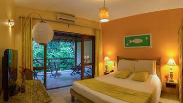 A hotel room with a bed, bedside lamps, a TV, a table with a chair, and a view of greenery through the door.