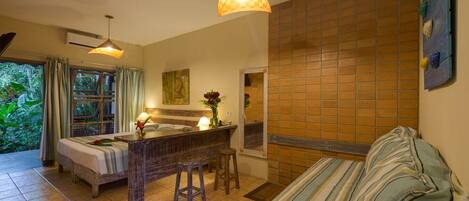 A room with a bed, a small bar area, a sofa, and a view of greenery outside.