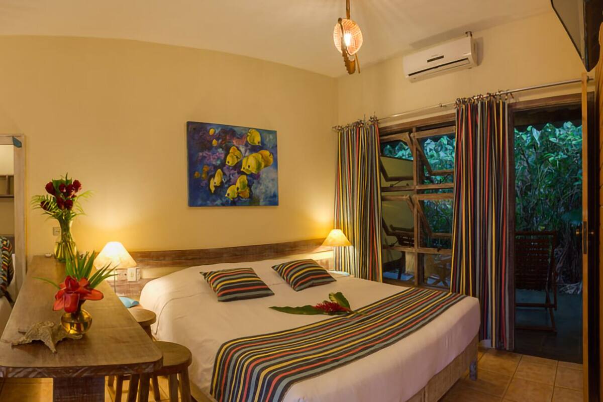 A hotel room with two single beds, a wooden table, a vase with flowers, and a large window with curtains.