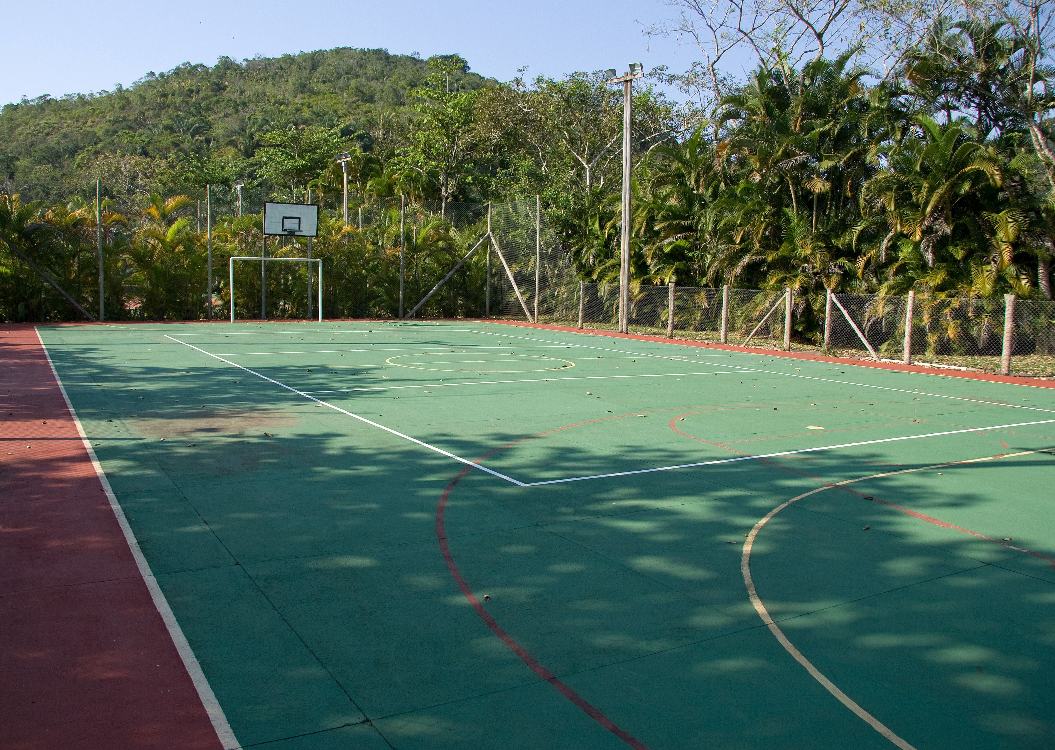 Una cancha de baloncesto al aire libre con superficie verde, rodeada por una valla y árboles.