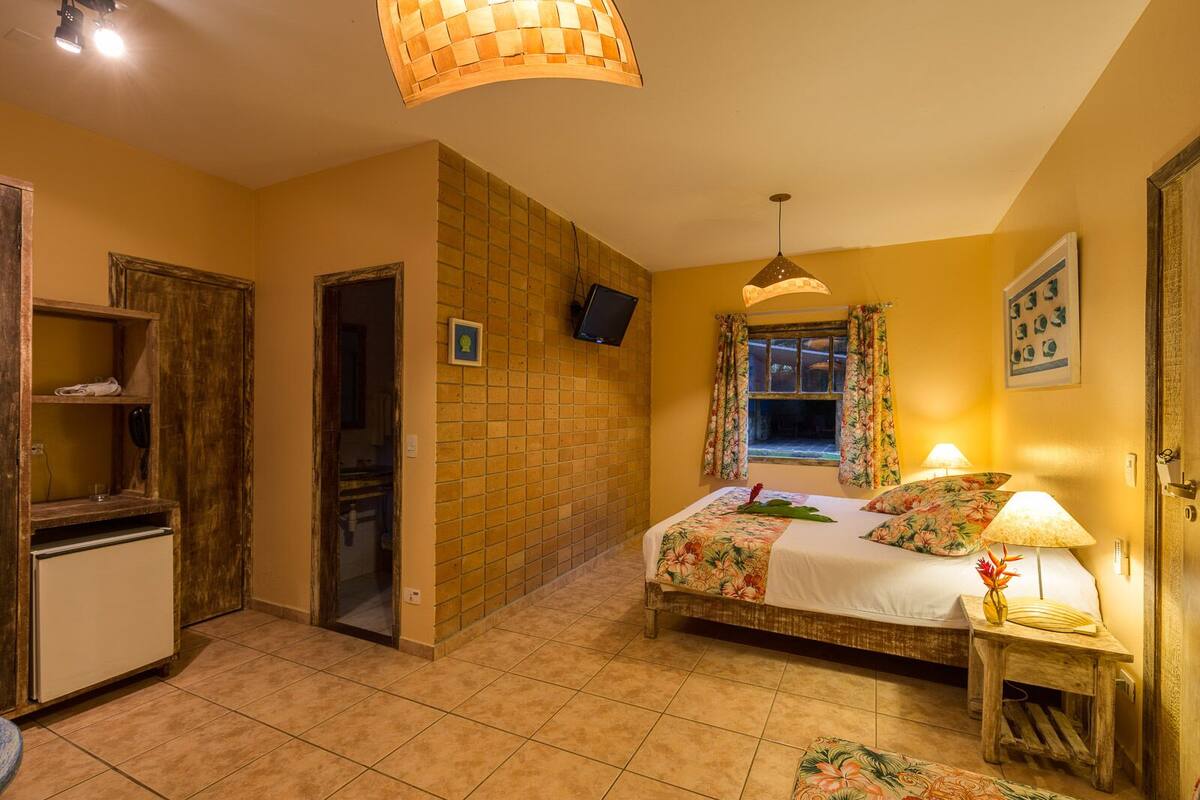 A bedroom with a bed, bedside lamps, a TV, a small table with a vase, and a door leading to another room.