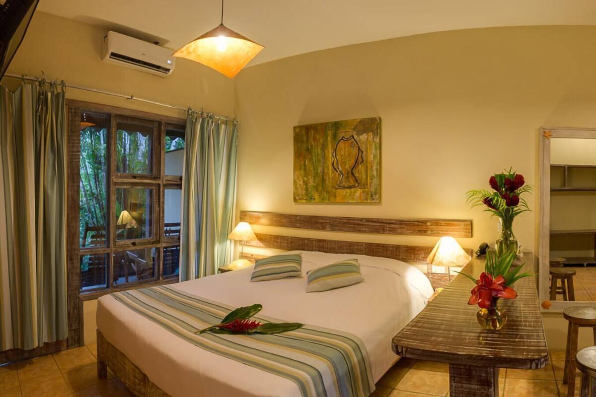 A hotel room with a bed, a sofa, a small table, a mirror, and a window with curtains.