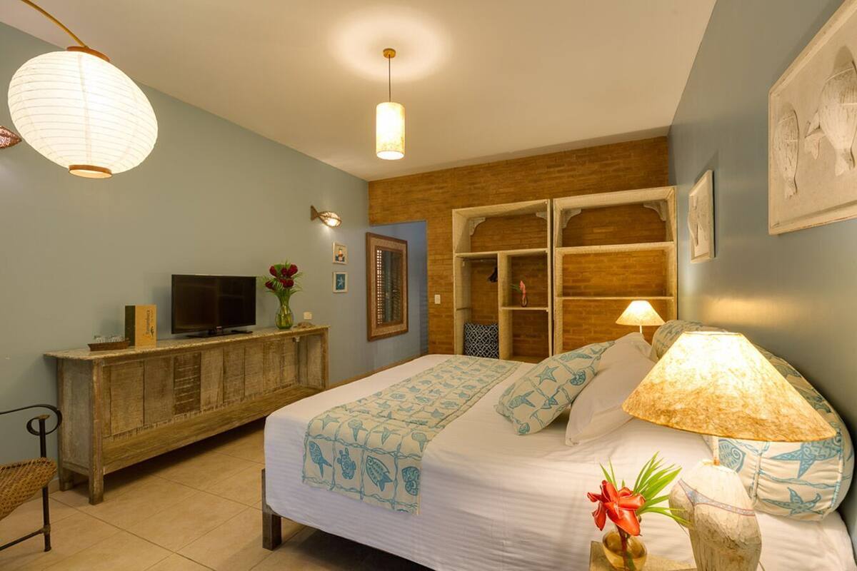 A bedroom with a bed, bedside lamp, TV, wooden dresser, and a wall-mounted shelf.