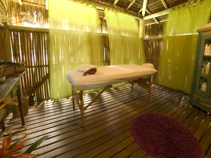 Una habitación con piso de madera, una cama con colchón blanco y cabecera marrón, una cortina verde y un mueble de madera con botellas.