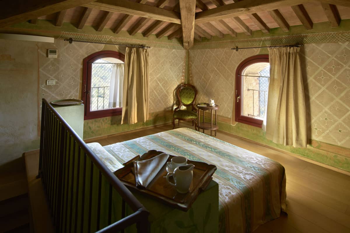 A room with a bed, a chair, a small table, and two windows with curtains.