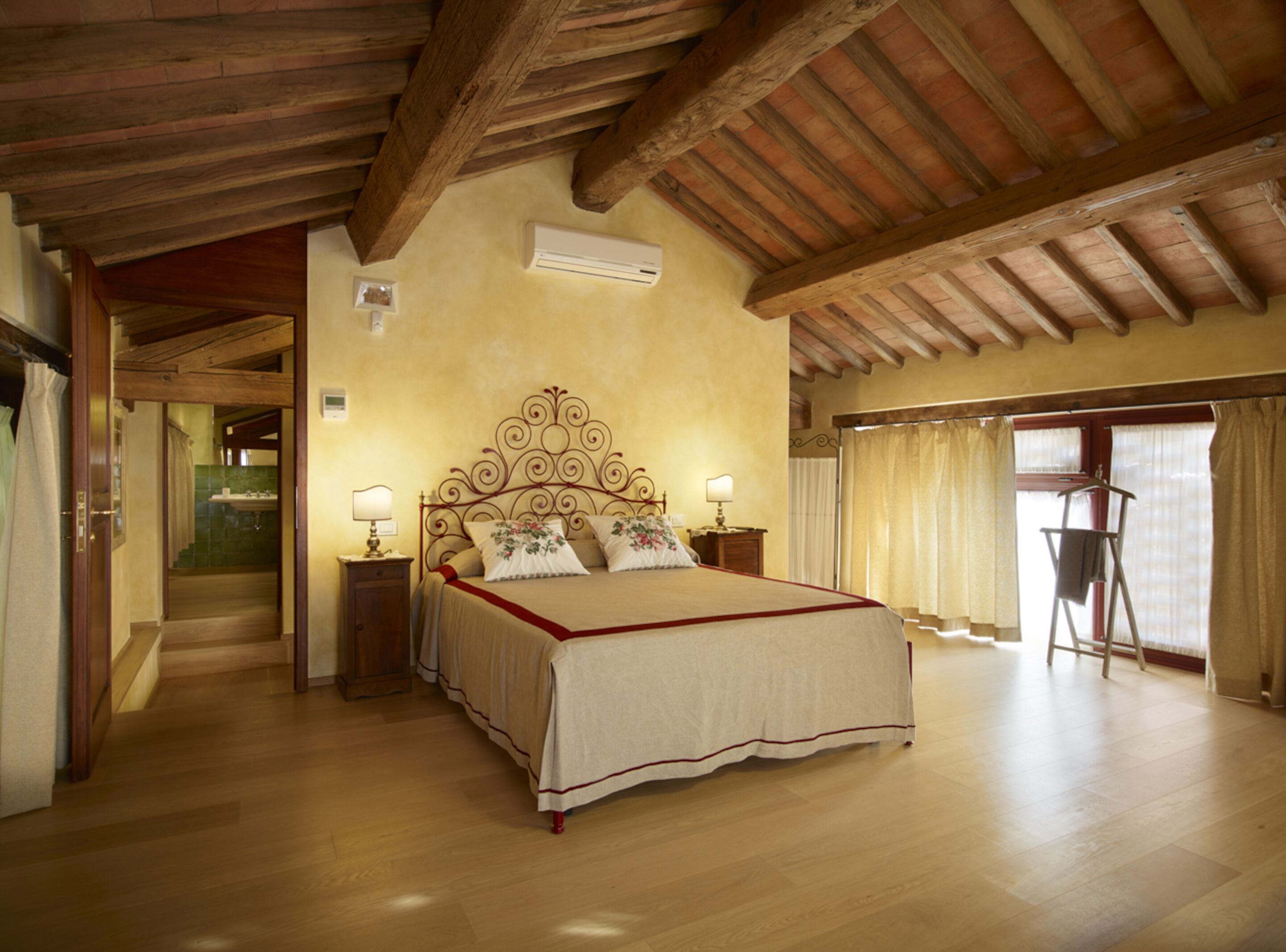Un dormitorio espacioso con una cama grande, vigas de madera en el techo, una mesita de noche pequeña con una lámpara y una ventana con cortinas.