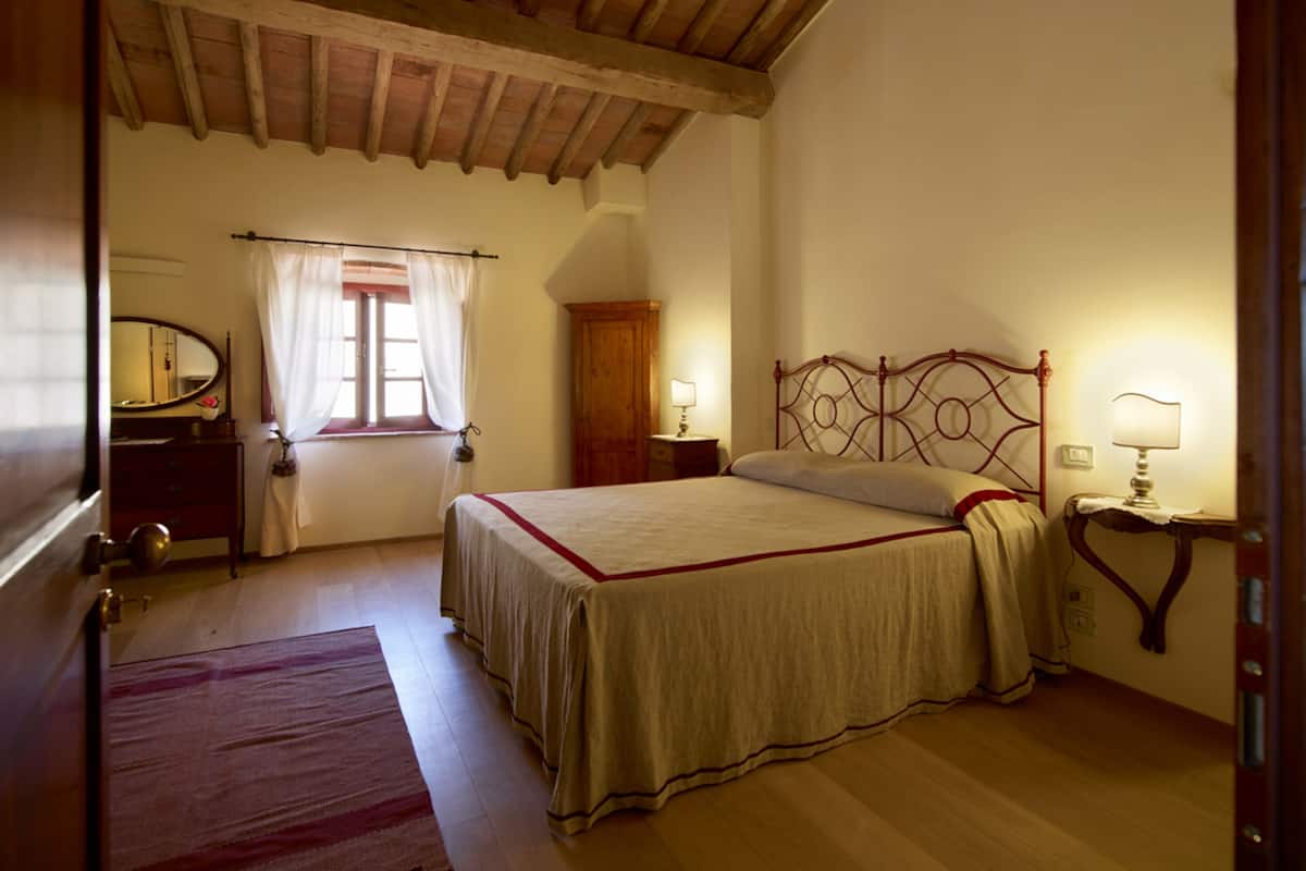 A bedroom with a wooden ceiling, a bed with a headboard, a nightstand with a lamp, a dresser, a mirror, and a window with curtains.