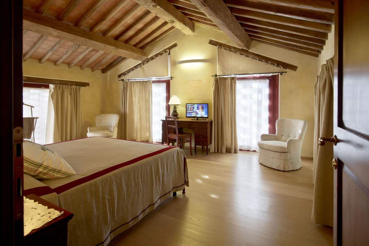 A bedroom with a large bed, two chairs, a desk with a TV, and wooden beams on the ceiling.