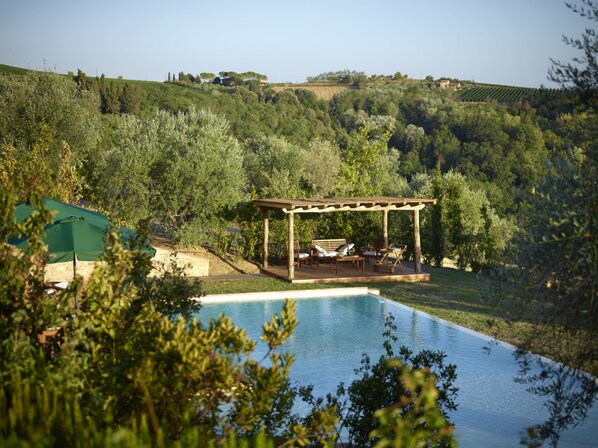 A pool with a wooden deck and lounge chairs, surrounded by lush greenery and hills in the background. - B&B Borgo Petrognano - Residenza d'epoca (Barberino Tavarnelle)