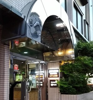 Entrada de un edificio con puerta de vidrio, toldo metálico y un cartel.